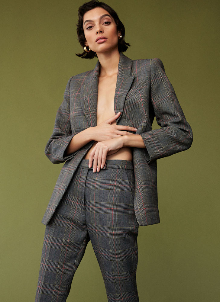 A woman in a grey plaid suit stands against an olive background. She is fair-skinned with short brown hair and gold hoop earrings.