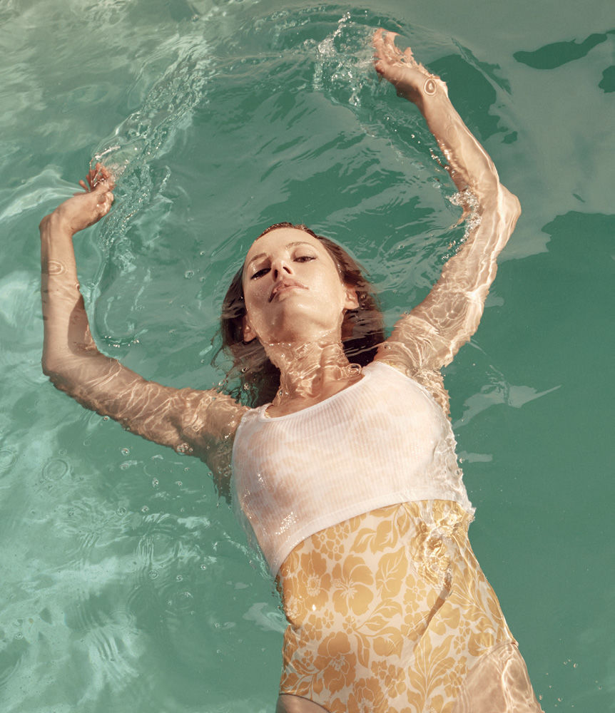 Woman floating in a pool of turquoise water with her arms raised above her head. She is wearing a floral one-piece swimsuit.