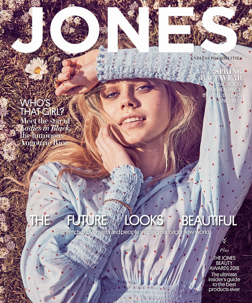Fashion magazine cover featuring a blonde woman in light blue polka dot dress, lying in a field of grass and white flowers with hand on her face.