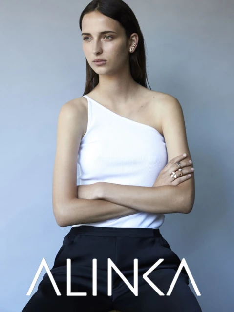 Woman in a white top and dark pants sits with arms crossed, looking to the left, showcasing ear and hand jewelry against a grey background.