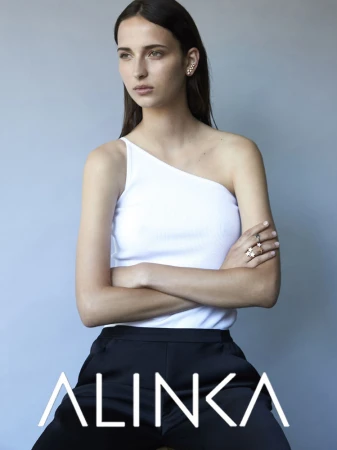 Model with arms crossed wearing a white one-shoulder top and dark trousers against a grey backdrop