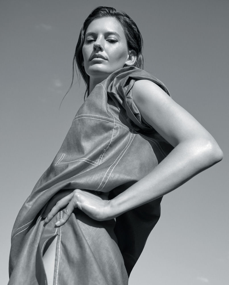 Monochrome portrait of a stylish woman in an oversized, draped top, posing against a clear sky with a confident gaze and hand on hip.