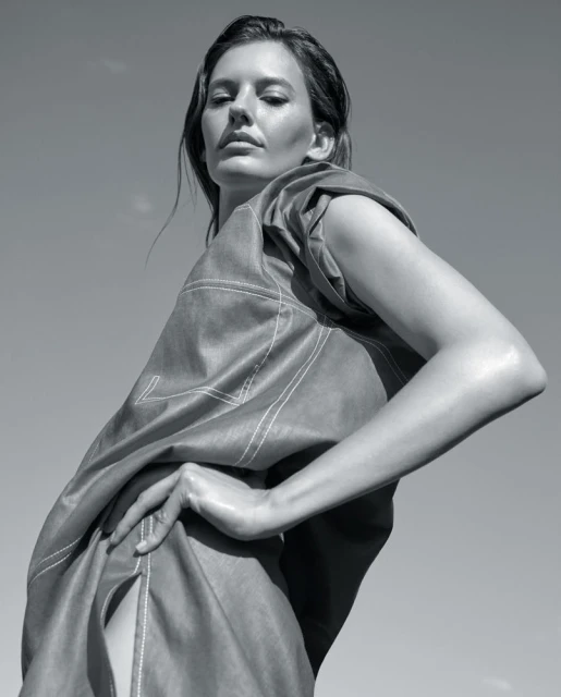 Monochrome portrait of a stylish woman in an oversized, draped top, posing against a clear sky with a confident gaze and hand on hip.