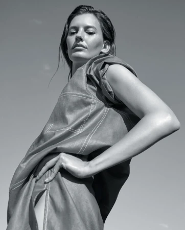 Monochrome portrait of a stylish woman in an oversized, draped top, posing against a clear sky with a confident gaze and hand on hip.