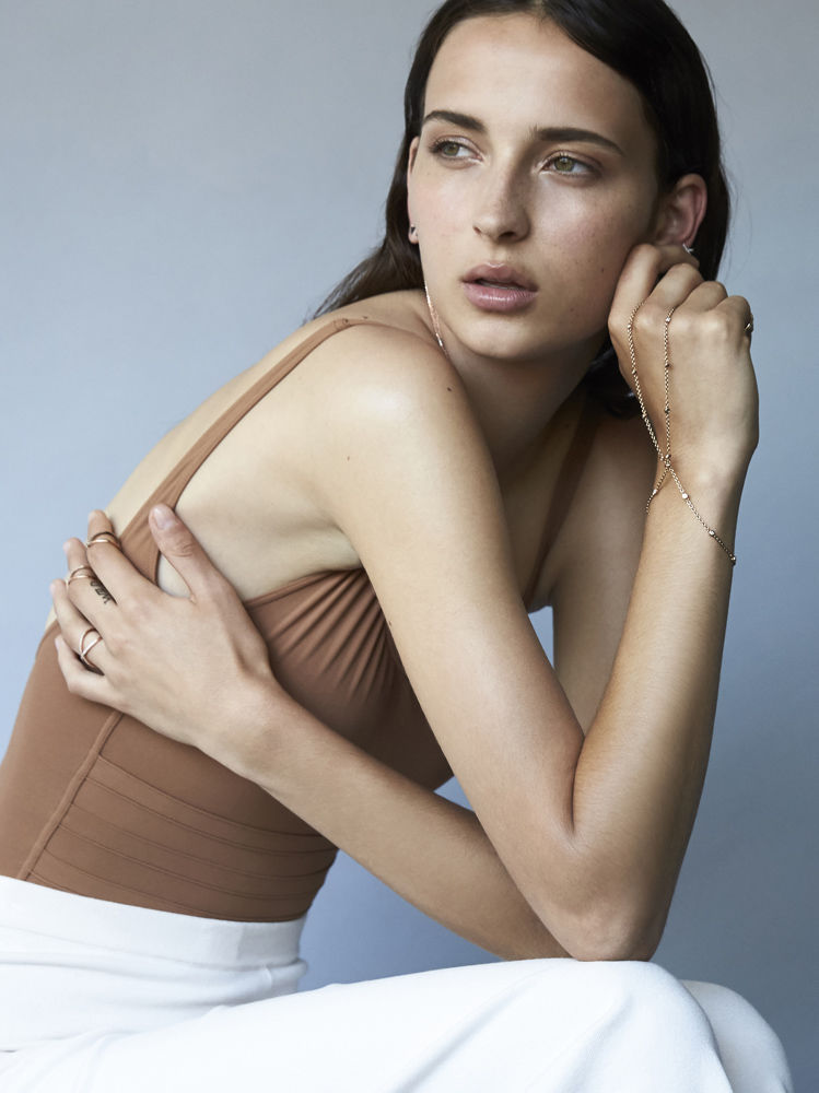 Portrait of an adult woman in a brown top and white pants in a photo studio, posed against a grey background, showcasing jewelry.
