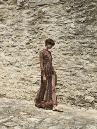 Model with short dark hair wearing a patterned long dress with fringe walking in front of a stone wall