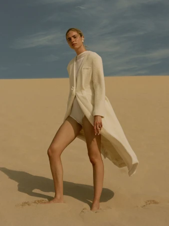 Model standing barefoot on a sandy dune wearing a long cream coat and white leotard under an overcast sky.