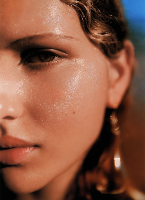 Close-up portrait of a model with wet-look skin, a mole, and gold dangling earrings against a blurred background.