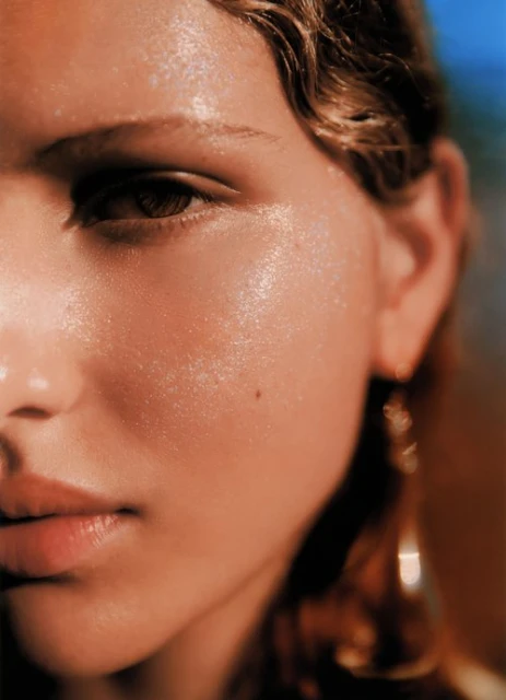 Close-up portrait of a model with wet-look skin, a mole, and gold dangling earrings against a blurred background.