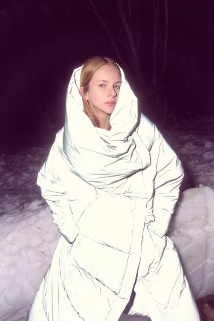 Woman wearing reflective coat stands in snow at night. The image features dark, snowy environment with an interesting play of light and shadow.
