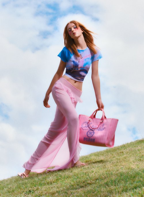 Young woman walks in grass with pink purse and see-through pants against a cloudy sky, fashion and summer vibes, outdoors, style.