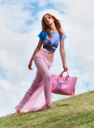 Red-haired model in blue graphic t-shirt and pink sheer trousers holding pink handbag on grassy hill against cloudy sky