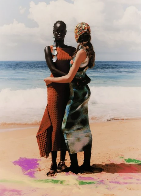 Two women embrace on a sandy beach with ocean waves and cloudy sky backdrop, wearing colorful dresses and standing in vibrant, patterned sand.