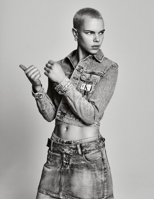 A person with a buzzcut poses in a denim jacket and skirt in a studio setting with a neutral background, looking over their shoulder.