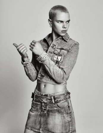 A person with a buzzcut poses in a denim jacket and skirt in a studio setting with a neutral background, looking over their shoulder.