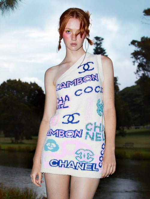 Young woman in a one-shoulder Chanel-branded dress stands outdoors with a lake and trees behind her against a cloudy sky.