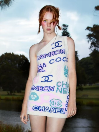 Young woman in a one-shoulder Chanel-branded dress stands outdoors with a lake and trees behind her against a cloudy sky.