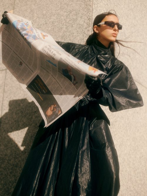 A woman in a black dress and sunglasses stands outside holding a newspaper with an editorial fashion style against a neutral background.