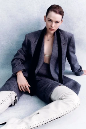 Woman with slicked-back hair wearing a grey blazer, trousers, and tall white lace-up boots seated against a pale wall.