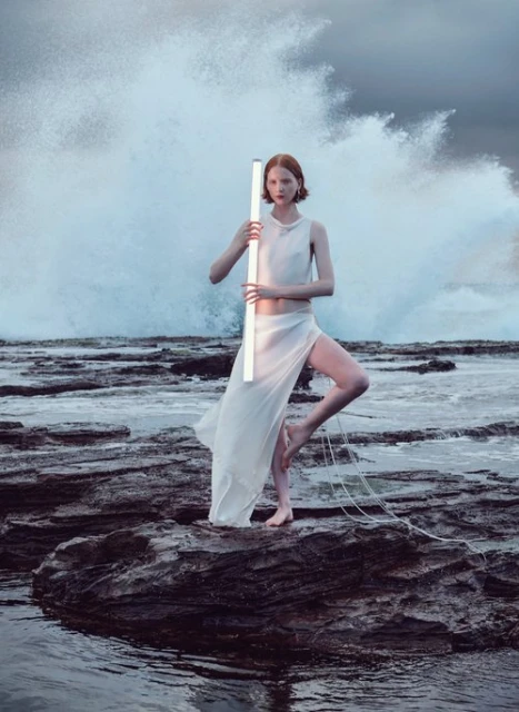 A woman in a white dress holding a light stands on rocks with a turbulent ocean backdrop, exuding artistic fashion and serene strength.