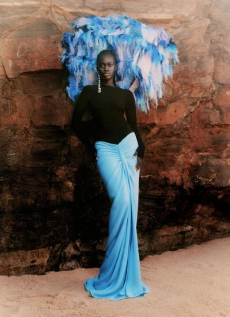 An adult female model poses in a long, light blue skirt with a black top, standing against a rocky backdrop with a feathered headpiece.