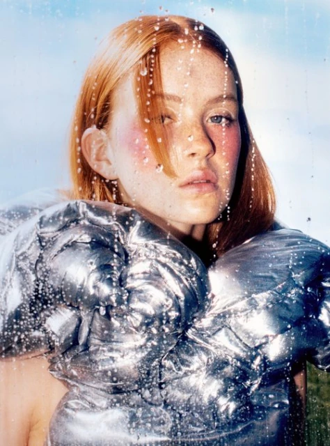 Close-up portrait of a red-haired woman with blush makeup and a metallic quilted top, captured against a water-streaked glass surface.