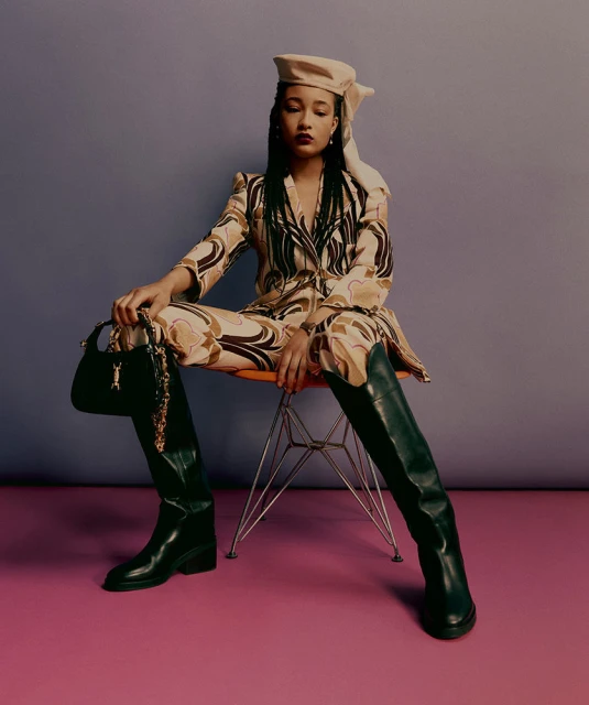 Young woman wearing a patterned suit with high boots sitting on an orange chair in a studio setting with a purse in her hand.