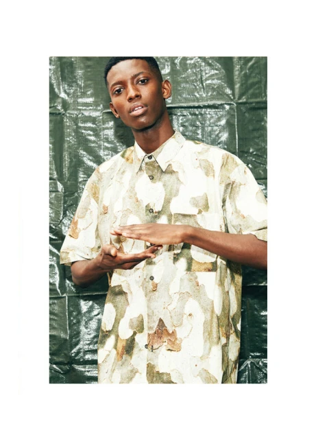 Portrait of a young adult Black male wearing a short-sleeve shirt with a natural pattern, posing indoors against a green textured backdrop.