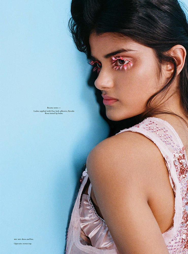 Close-up beauty shot of a young woman wearing a pink dress and flower petal eyelashes against a light blue backdrop in a studio.