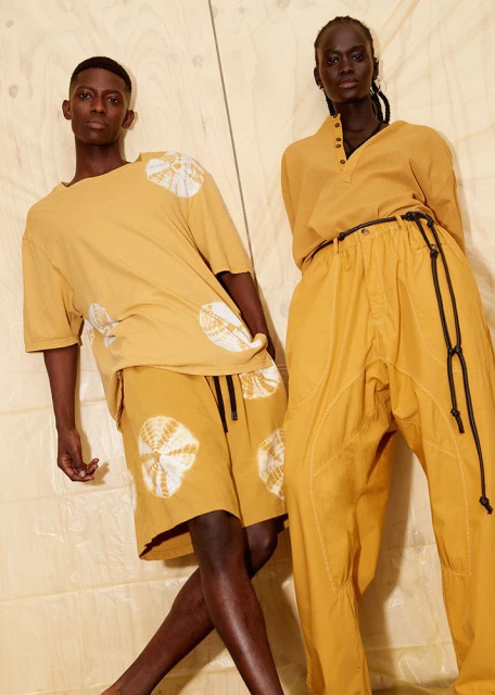 Two fashion models in mustard-yellow outfits pose against a plywood wall, showcasing a casual, stylish aesthetic with unique circle patterns.
