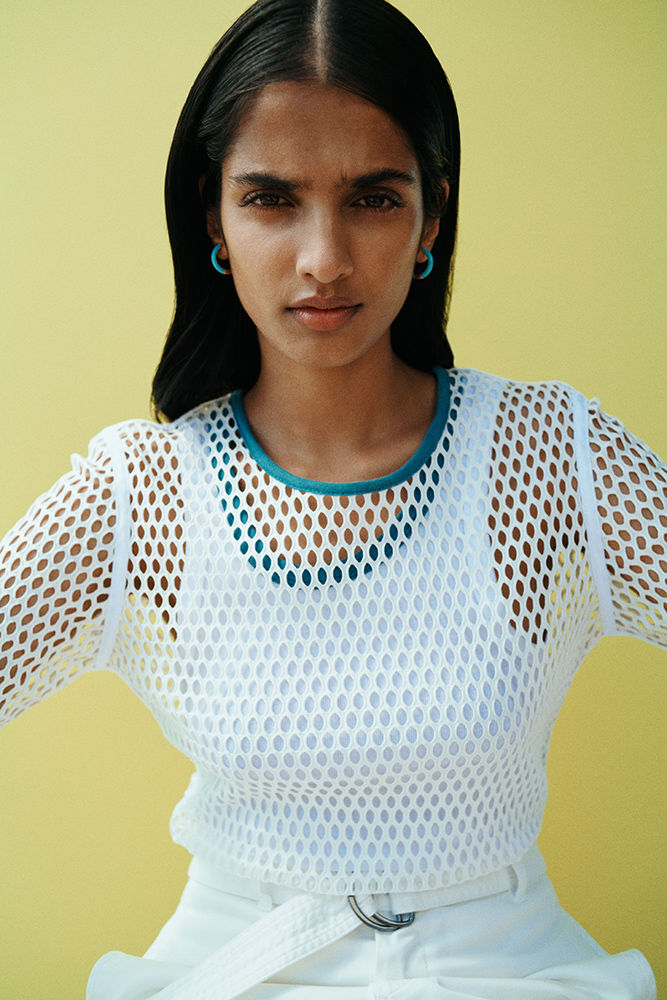 Portrait of a woman with dark hair wearing a white mesh top and white pants against a bright yellow background in a studio setting.