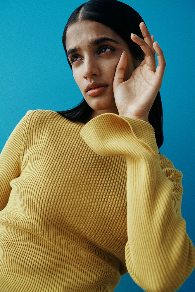 Close-up portrait of a woman in a yellow ribbed knit sweater with her hand raised near her face against a bright blue background