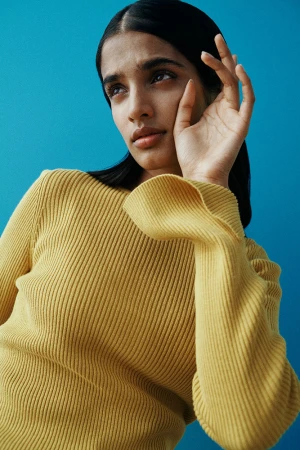 Close-up portrait of a woman in a yellow ribbed knit sweater with her hand raised near her face against a bright blue background