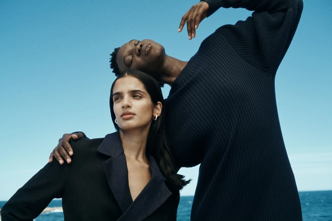 Two female models posing outdoors against a clear blue sky and ocean backdrop with make-up by Joel Babicci