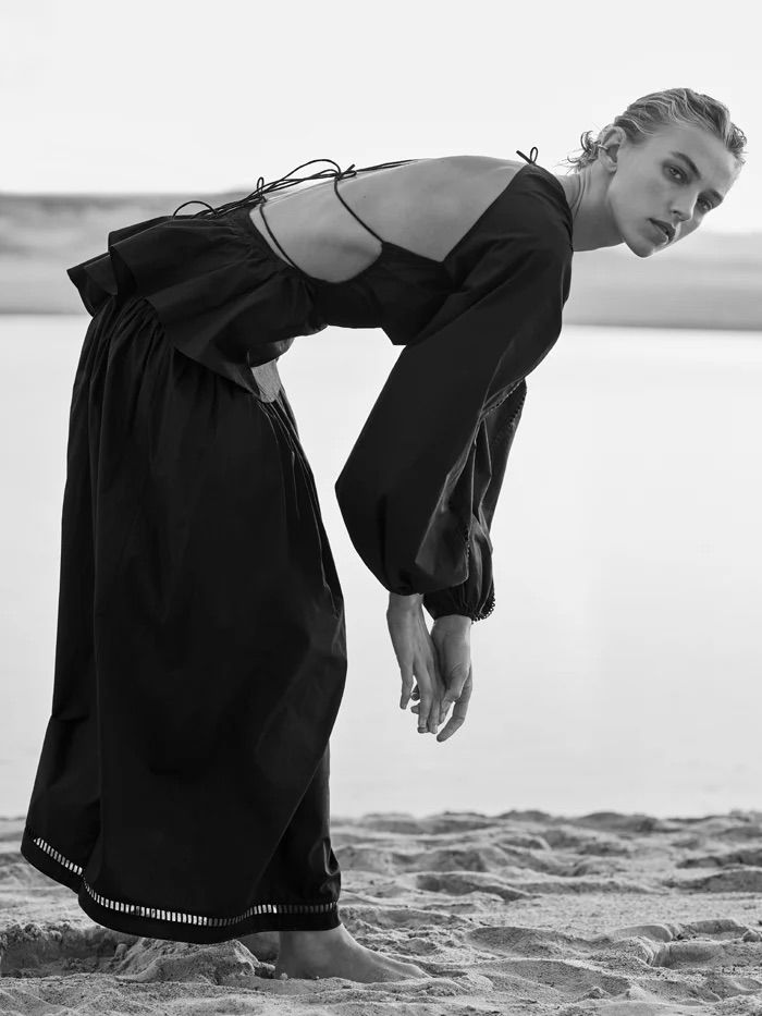 Model in a black dress bending forward on a sandy beach under an overcast sky by Joel Babicci