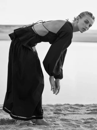 Model in a black dress bending forward on a sandy beach under an overcast sky by Joel Babicci