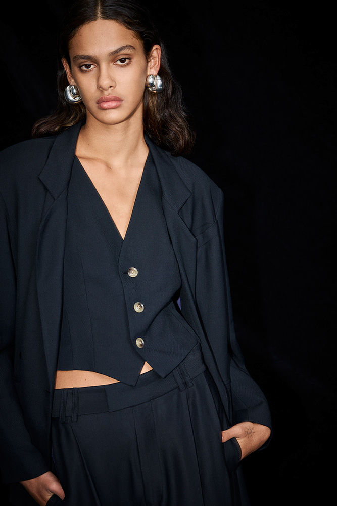 Fashion portrait of a woman wearing a navy suit with a vest and silver hoop earrings against a dark background.