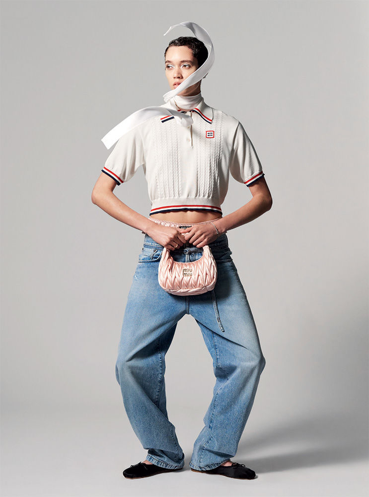 Fashion shot of a slender woman in an off-white top, jeans, and black flats, holding a pink Miu Miu handbag against a grey background.