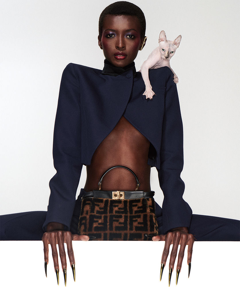 Elegant dark-skinned woman in a cropped navy blazer poses with a purse and a Sphynx cat on a white background in a fashion editorial.