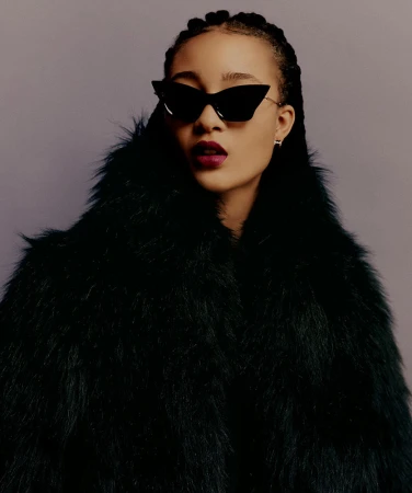 Model wearing dark cat-eye sunglasses and a dark furry coat with plum lipstick against a grey background