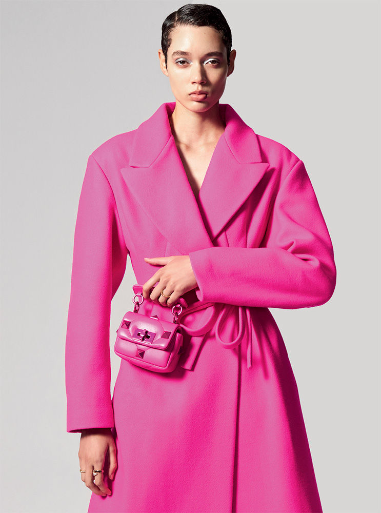 Woman in a bright pink coat and matching mini bag poses in a studio. The high fashion shot captures a beauty and style aesthetic.