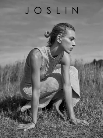 Monochrome fashion portrait of a female model crouching in a grassy field wearing a ribbed knit dress, with makeup by Joel Babicci.