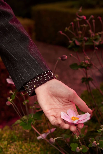 Close-up of a woman's hand gently holding a pink flower outdoors, highlighting the jacket cuff with decorative studs.