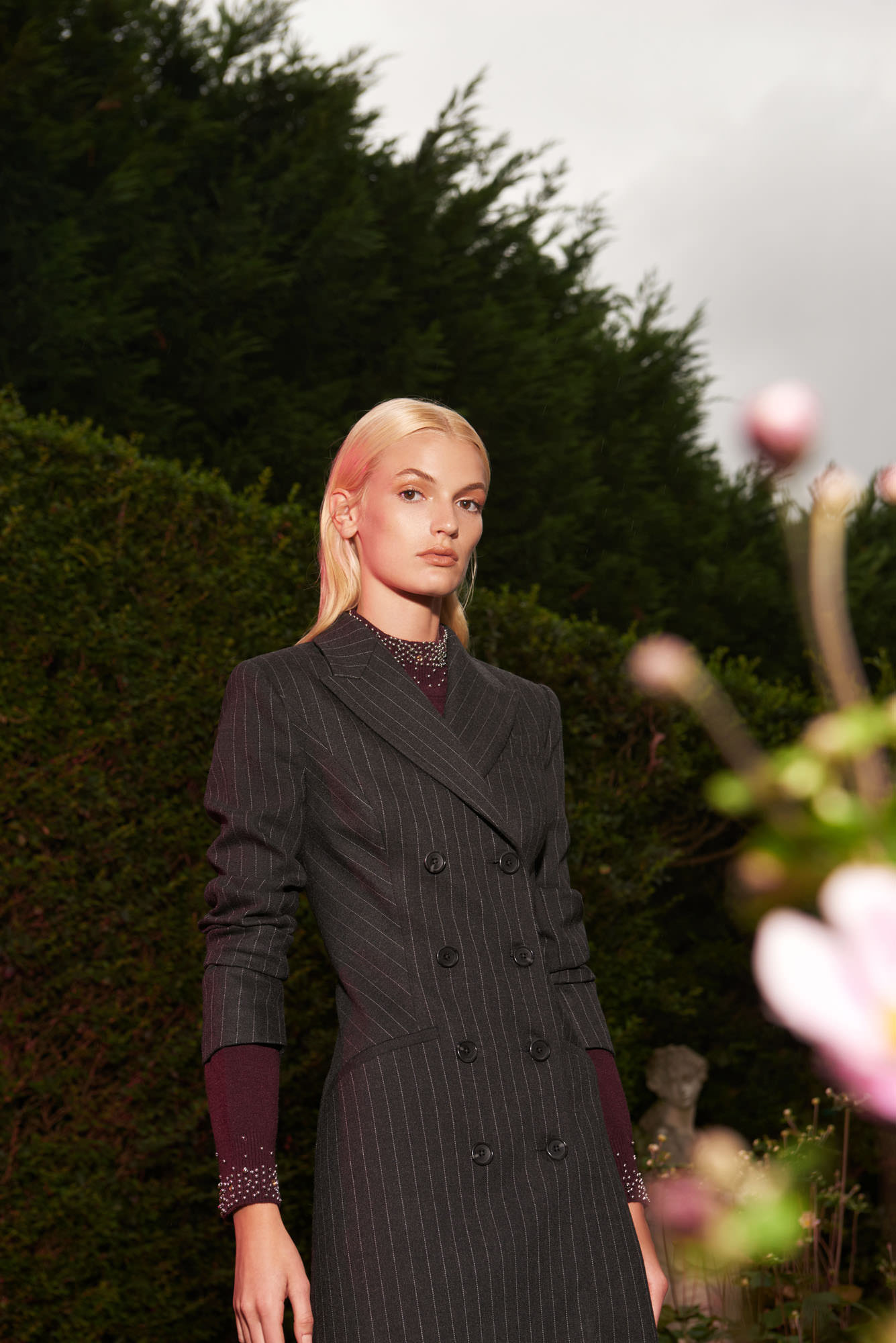 A woman in a pinstripe dress poses in an outdoor garden setting with green foliage and pink flowers in the foreground.