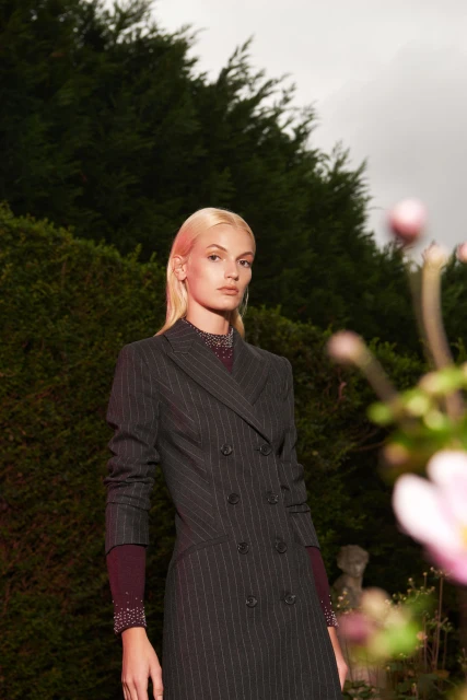 A woman in a pinstripe dress poses in an outdoor garden setting with green foliage and pink flowers in the foreground.