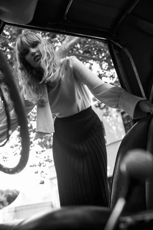 Monochrome low angle shot of a woman leaning into a car interior wearing a long sleeve blouse and ribbed skirt.
