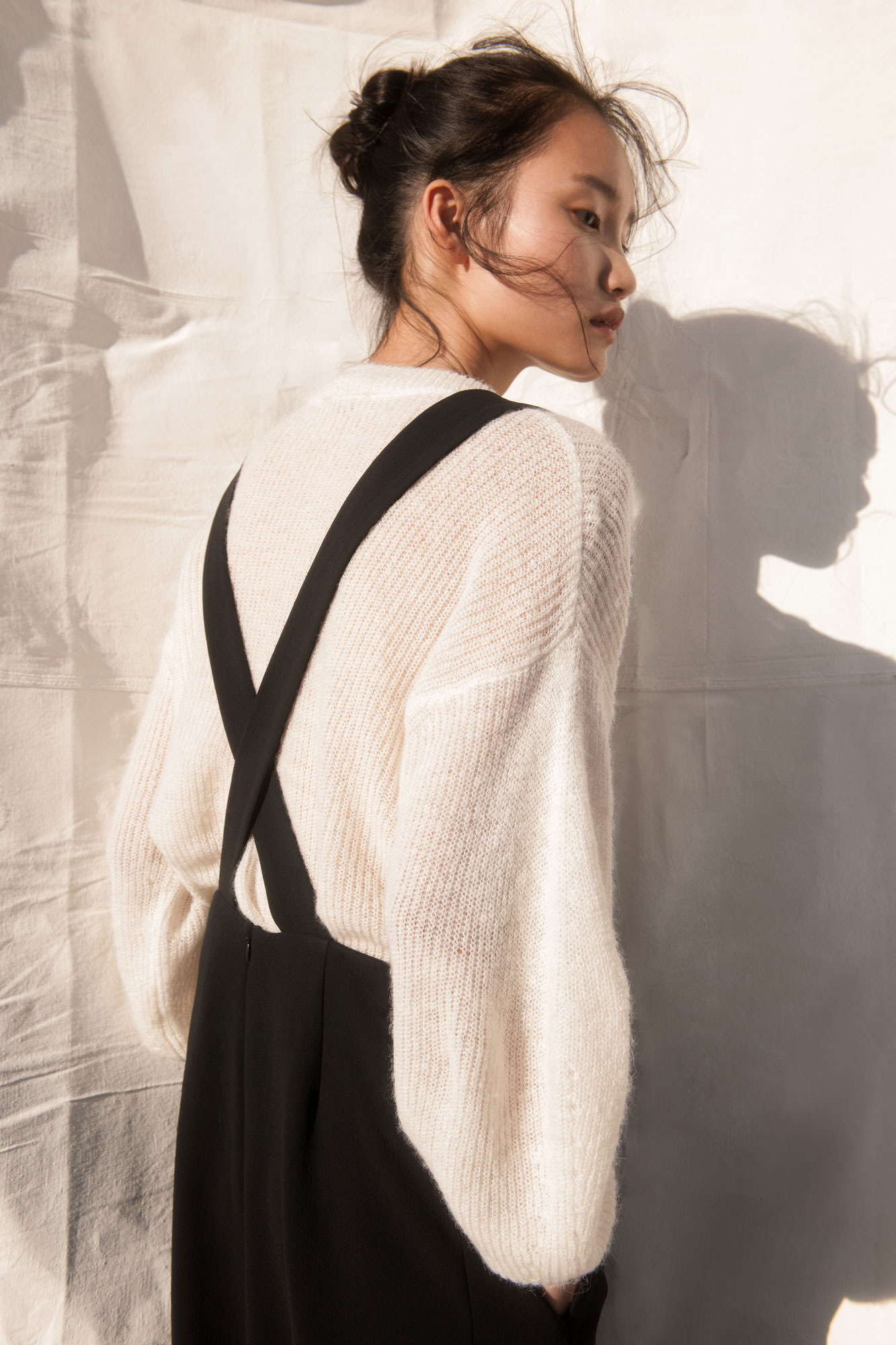 Portrait of a young Asian woman wearing a cream sweater and black overall dress posing against a white background with soft shadow.