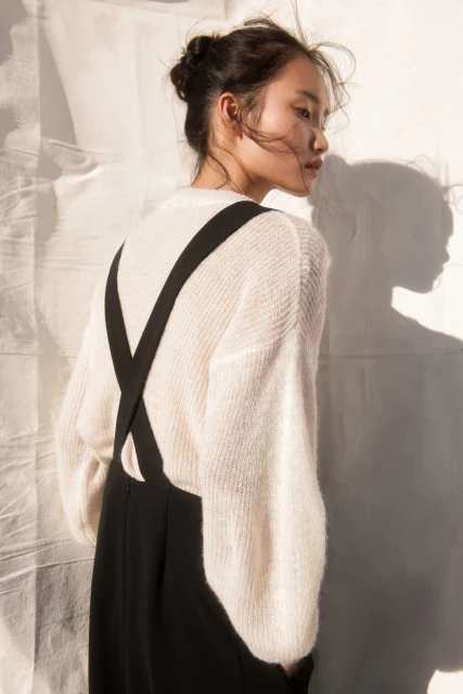 Portrait of a young Asian woman wearing a cream sweater and black overall dress posing against a white background with soft shadow.