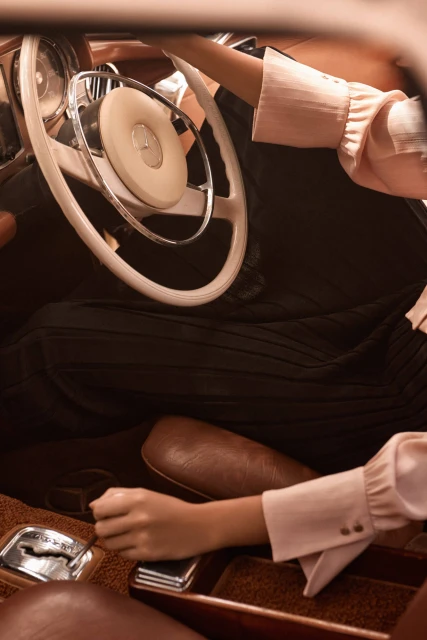 Close-up of a person driving a classic car with a white steering wheel, brown interior, and a hand on the gear shift.