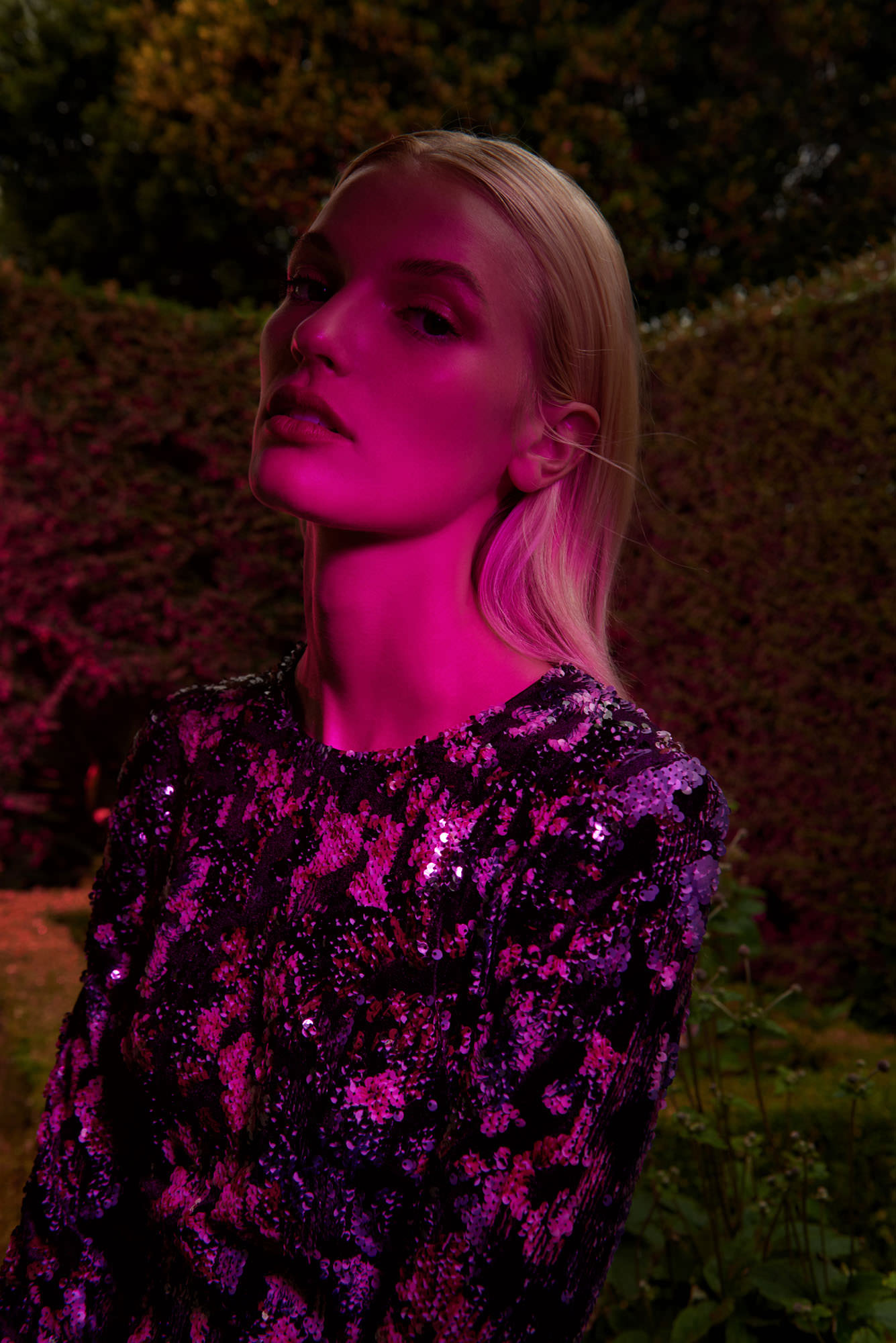 Portrait of a blonde woman wearing a sequined top illuminated by vibrant magenta lighting in an outdoor garden.